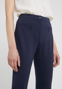 Femme portant un pantalon bleu marine avec des poches avant zippées et un haut blanc sans manches sur un fond uni.