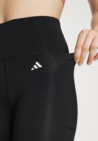 Black athletic leggings with a high waistband, featuring a white logo and a side pocket. Smooth texture and fitted design for comfort.