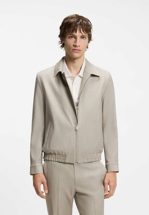 Junger Mann mit kurzem braunen Haar, der eine hellbeige Reißverschlussjacke über einem weißen Hemd mit Kragen und passenden beigen Hosen trägt.