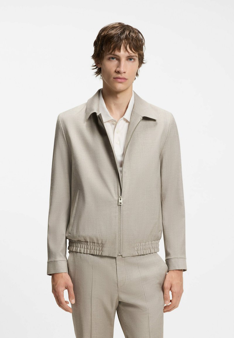 Jeune homme aux cheveux bruns courts portant une veste zippée beige clair sur une chemise blanche à col et un pantalon beige assorti.