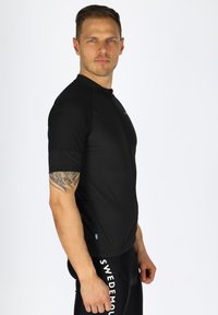Schwarzes Sportshirt mit Reißverschluss vorne, kurzen Ärmeln und körpernaher Passform. Mit schwedischem Flaggenmotiv am Saum. Glatte Textur.