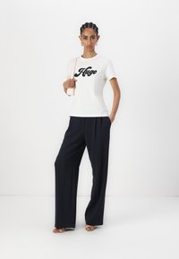 T-shirt en coton blanc avec logo "Hugo" noir, associé à un pantalon large taille haute bleu marine. Le mannequin tient un sac à bandoulière rose clair.