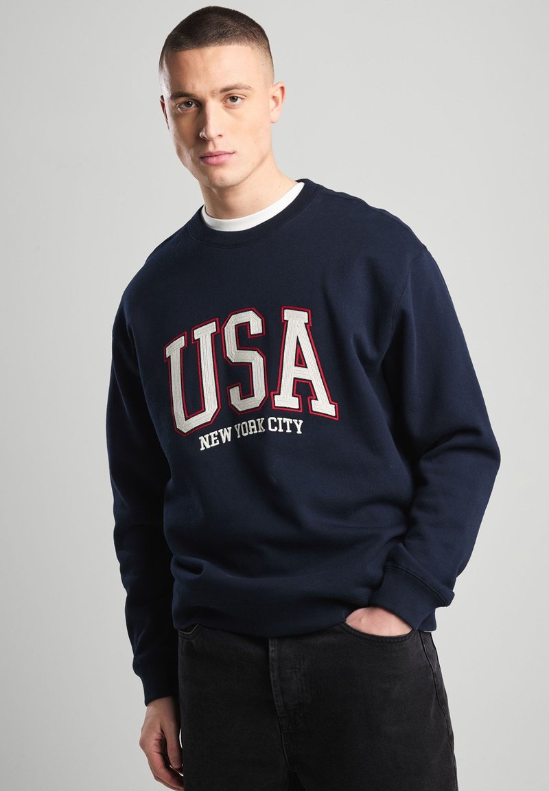 Navy sweatshirt featuring "USA" and "New York City" in bold red and white lettering; round neckline; ribbed cuffs; casual fit.