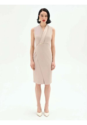 Donna con capelli neri corti che indossa un abito beige senza maniche e tacchi bianchi, in piedi davanti a uno sfondo bianco semplice.