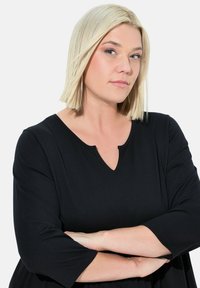 Černé šaty s výstřihem do V a třičtvrtinovými rukávy, volný střih a hladká textura. Modelka se světlými blond vlasy pózuje se založenýma rukama.