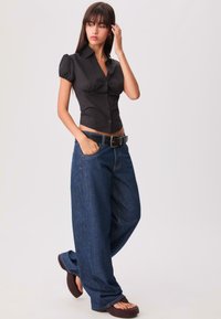Zwarte getailleerde blouse met pofmouwen en knopen, gecombineerd met losse blauwe denim jeans en een zwarte riem. Bruine schoenen maken de outfit af.