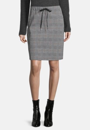 Vrouw draagt een grijze geruite rok tot op de knie met een zwarte koordsluiting, een zwarte trui met lange mouwen en zwarte glanzende enkellaarzen.