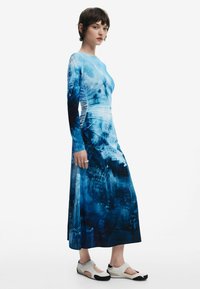 Maxi haljina s printom plavog urbanog pejzaža, dugim rukavima, uskim tijelom i lepršavom suknjom. U kombinaciji s bijelim cipelama s crnim detaljima.