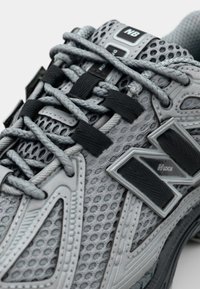 Harmaat urheilukengät, joissa on verkko-osia, mustat nauhat ja näkyvä harmaa "N"-logo. Varustettu tekstuurilla ja tukevilla päällysteillä.