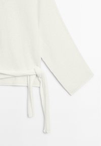 White knit top featuring long sleeves and a side knot detail, made from soft, textured fabric with a relaxed fit.