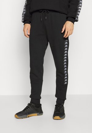 Mężczyzna ubrany w czarne spodnie dresowe z białymi paskami z logo po bokach oraz czarne trampki z gumowymi podeszwami, stojący na białym tle.