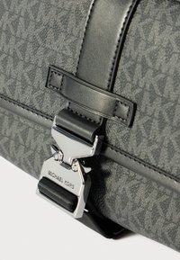 Primo piano di una borsa grigia Michael Kors con motivo testurizzato del logo MK e una fibbia di chiusura argentata incisa con il nome del marchio.