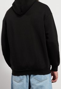 Hombre visto de espaldas vistiendo una sudadera negra con capucha y jeans de mezclilla azul claro frente a un fondo blanco liso.