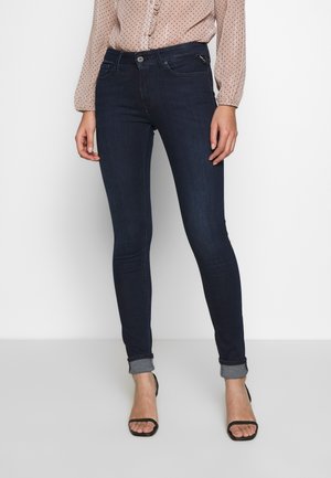 Femme portant un jean skinny bleu foncé avec des revers, une blouse rose clair à petits pois noirs et des talons ouverts noirs, debout devant un fond uni.