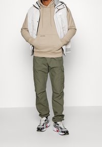 Chaleco acolchado blanco sobre una sudadera beige, combinado con pantalones cargo verde oliva y zapatillas deportivas multicolores con detalles en negro, gris y rojo.