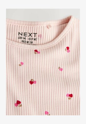 Svetlo roza rebrasta majica s malimi srčastimi motivi v rdeči, roza in rjavi barvi. Etiketa prikazuje ime blagovne znamke in navodila za nego.