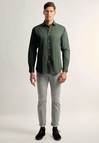 Camisa verde de botones con mangas largas, combinada con jeans gris claro, con los puños doblados, y zapatos oscuros. Tela suave, corte clásico.