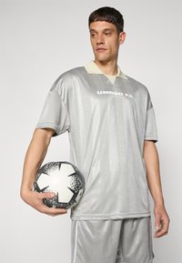 Grå fotballdrakt med lys krage, med et geometrisk mønster og teksten "UMBRO F.C.". Holder en svart og hvit mønstret fotball.