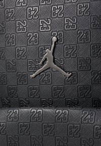 Juodos tekstūruotos medžiagos, kurioje yra iškilūs „23“ ir „23“ logotipų raštai. Centre išryškintas sidabrinis Jumpman logotipas.
