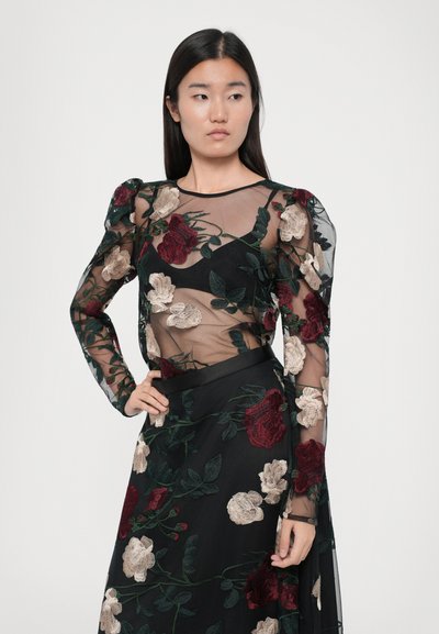 Zwarte doorschijnende jurk met lange mouwen, met een gemêleerd bloemenborduursel in rood, crème en groen. Model poseert met handen op de heupen.