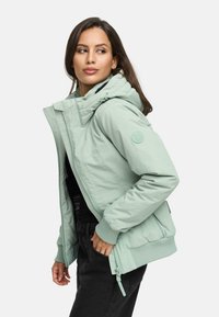 Light green puffer jacket with a hood, featuring a zip front, ribbed cuffs, and logo patch on the sleeve. Worn over a black top.
