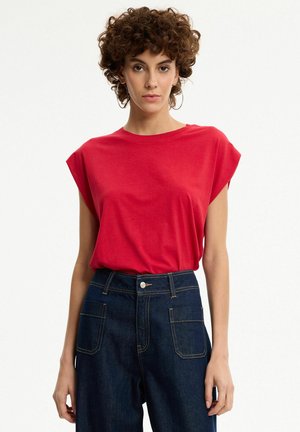 Mujer con cabello rizado y castaño que lleva una camiseta roja sin mangas y vaqueros azul oscuro de talle alto con bolsillos delanteros, posando frente a un fondo blanco.