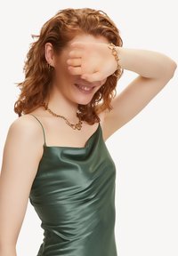 Groene satijnen jurk met een gedrapeerd ontwerp, gecombineerd met een gouden ketting en armband. Haar is krullend en schouderlang. Hand voor het gezicht.