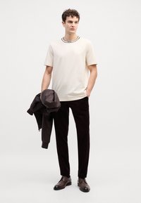T-shirt beige avec détail noir au col, pantalon en velours côtelé foncé, chaussures en cuir marron, tenant une veste sombre. Fond uni.