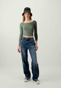 Green long-sleeve crop top, blue ripped jeans, black bucket hat, and black low-top sneakers. Slim fit and casual style.