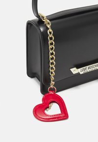 Borsa nera con finitura liscia, dotata di una catena dorata e di un ciondolo a forma di cuore rosso. Include una placca con il logo sul davanti.