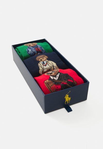 Ceturto pāru zeķu zils kastē: zaļas ar lāci vestē, jūras zila ar lāci džemperī, sarkanas ar lāci mētelī, adīta tekstūra.