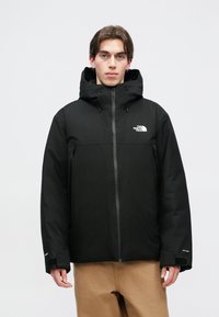 Zwarte geïsoleerde jas met een capuchon, logo op de borst, voorritsen en klittenbandmanchetten. Gemaakt van waterafstotend materiaal.