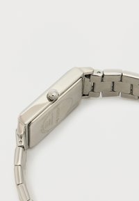 Hõbedane metallkell, millel on ristkülikukujuline nähik, metallist rihm, tekstuuriga nupud külgedel ja graveeritud bränding tagaküljel.