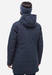 Veste imperméable navy avec capuche, coupe longue et texture lisse. Comprend des poignets ajustables et un logo de marque discret sur la manche.