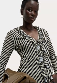 Donna che indossa una blusa a strisce verticali bianche e nere con volant, mentre tiene una borsa di pelle marrone su uno sfondo chiaro e uniforme.