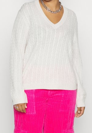 Personne portant un pull blanc en maille torsadée à col en V et un pantalon en velours côtelé rose vif, accessoirisée d'un collier chaîne en argent et de bagues.