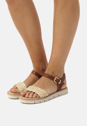 Sandalias de cuero marrón con tiras de paja trenzadas, suela acolchada y hebilla ajustable en el tobillo, que presentan un acabado natural y texturizado.
