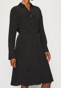 Abito camicia nero realizzato in tessuto morbido, con colletto, apertura con bottoni, maniche lunghe e una cintura coordinata in tessuto in vita.