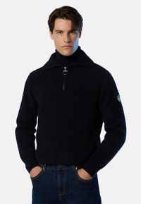 Ein gestrickter navyblauer Pullover mit Kragen zum Reißverschluss, geripptem Muster und tailliertem Design. Verfügt über einen runden Reißverschluss-Zipper und ein Logo am Ärmel.