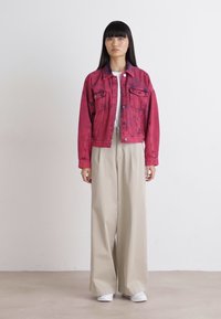 Giacca di jeans rossi con due tasche frontali, abbinata a pantaloni beige a gamba larga e sneakers bianche. Il modello è in piedi contro uno sfondo semplice.