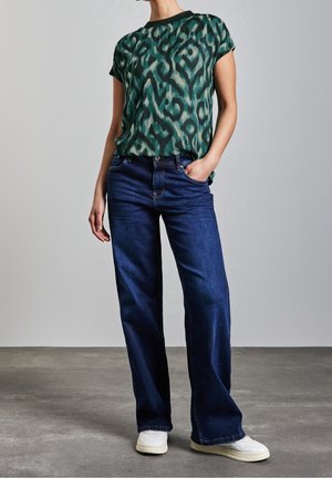 Person iført grøn mønstret kortærmet top, mørkeblå vidde jeans og hvide sneakers, stående med den ene hånd i lommen.