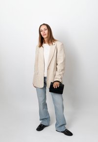 Femme en blazer beige, chemise blanche, jean bleu clair et mocassins noirs, tenant un petit sac à main noir, debout devant un fond blanc uni.