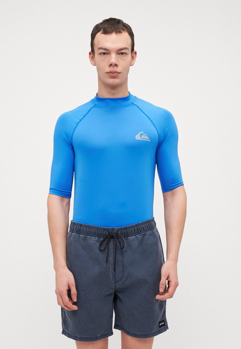 Blå kortermet rashguard med høy krage og Quiksilver-logo. Kombinert med grå badeshorts med elastisk midje og snøring.