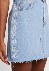 Světle modrá džínsová minisukně s bílou květinovou výšivkou po levé straně, ohrnutým lemem a klasickým designem s pěti kapsami.