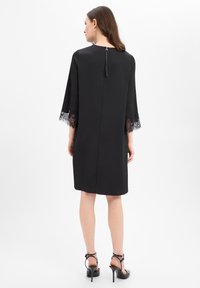 Zwarte jurk met driekwartse mouw van kant, gladde textuur, ritssluiting aan de achterkant, kniehoogte, eenvoudige silhouet, gedragen met zwarte sandalen met bandjes.