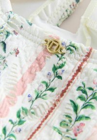Gros plan sur un tissu blanc avec une broderie florale verte et rose, des motifs texturés, et un petit fermoir décoratif doré avec des boucles en corde.