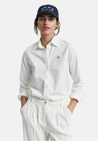 Weißes Hemd mit langen Ärmeln und Knopfleiste, mit einem kleinen Logo auf der Brust. Kombiniert mit weißen gestreiften Hosen und einer marineblauen Kappe mit Branding.