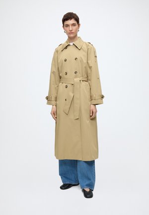 Άτομο με κοντά μαλλιά που φοράει μπεζ διπλής σειράς καμπαρντίνα, μπλε τζιν με φαρδιά μπατζάκια και μαύρα παπούτσια, στέκεται μπροστά σε λευκό φόντο.