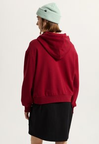 Red hooded sweatshirt with a relaxed fit, ribbed cuffs, and hem, worn over a black skirt. Accompanied by a light green beanie.