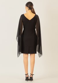 Schwarzes Kleid mit taillierter Silhouette, transparenten Fledermausärmeln, V-Ausschnitt hinten und knielangem Saum. Gestylt mit schwarzen Riemchen-High-Heels.
